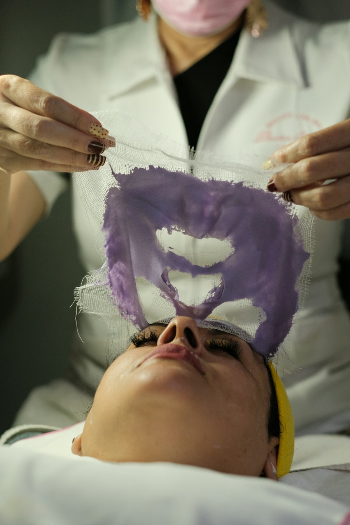 Esthetician removing a purple facial mask from a woman's face.